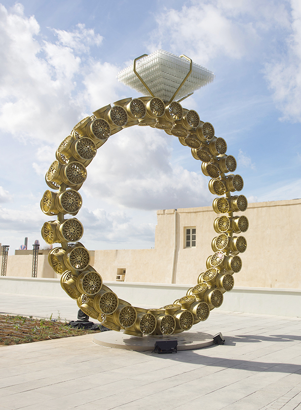 A large, outdoor sculpture of a ring made from recycled car parts. The band of the ring is composed of numerous golden car wheels, and the diamond is made of stacked plastic bottles.